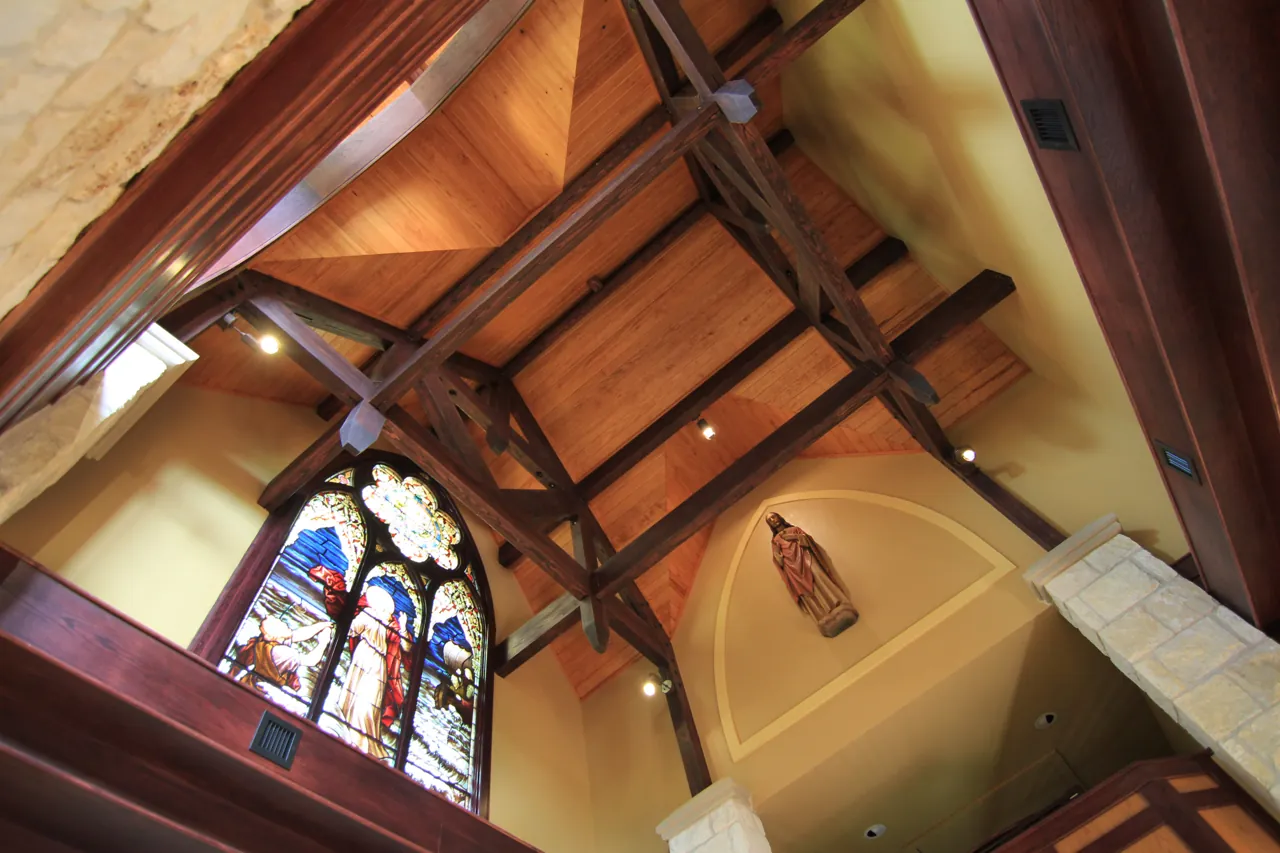 Dark-stained timber roof trusses and board ceiling over a limestone chapel, with a stained glass window