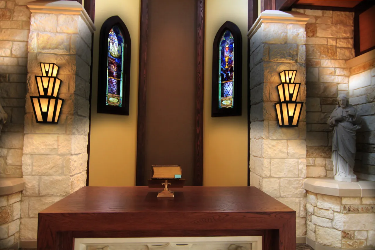 Wood altar before stained glass lancets, wall sconces and a statue in a limestone sanctuary