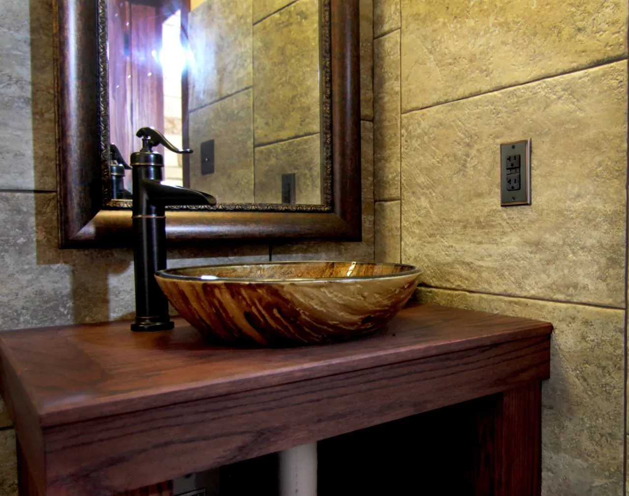 Dark wood vanity top with a glass vessel sink, bronze faucet and framed mirror against stone